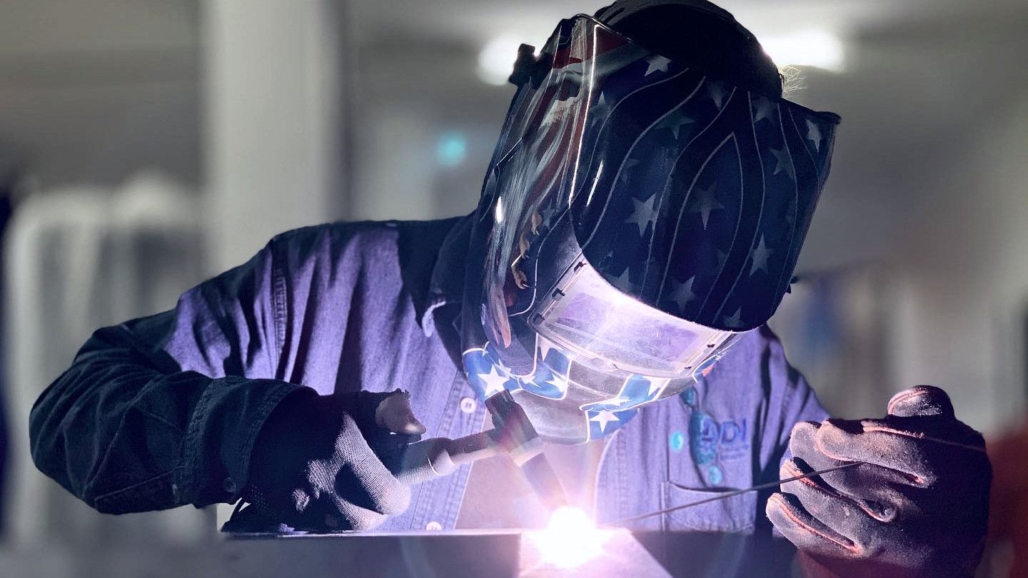 Welder in a DDI shirt working under a welding helmet, sparks flying
